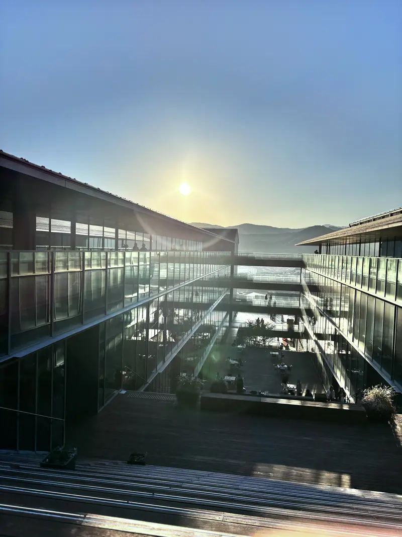 夕日に照らされる、ガラス張りのモダンな複合施設。ウッドデッキや中庭で人々がくつろいでいる。